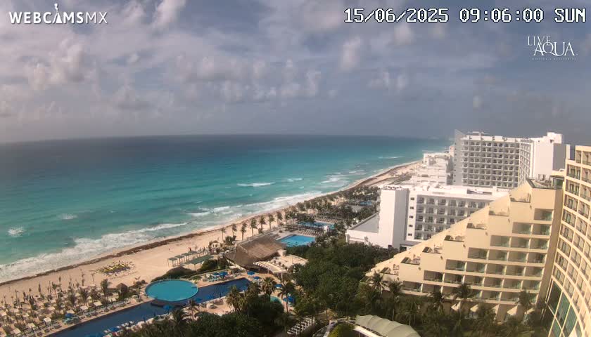 A partly cloudy sunny day overlooking a turquoise ocean with waves lapping a sandy beach lined with resorts and palm trees.