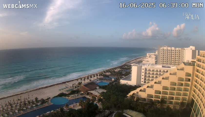 A partly cloudy morning shows a long stretch of beach and turquoise ocean next to a resort area with multiple hotels and swimming pools.