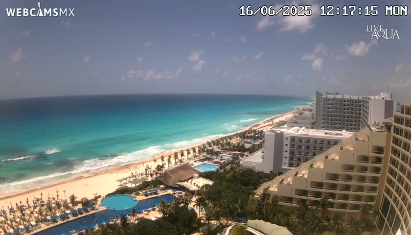 An aerial view shows a partly cloudy day over a long sandy beach with turquoise water, numerous resort hotels, and swimming pools.