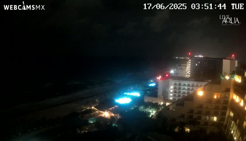 A nighttime view of a beach and several brightly lit hotels under a partly cloudy sky.