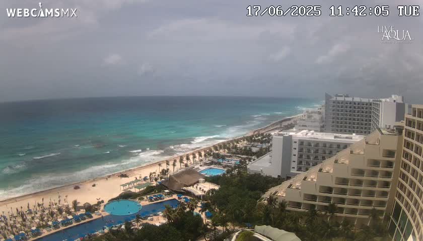 An ocean beach with turquoise water under an overcast sky is viewed from a high vantage point overlooking resort hotels and swimming pools.