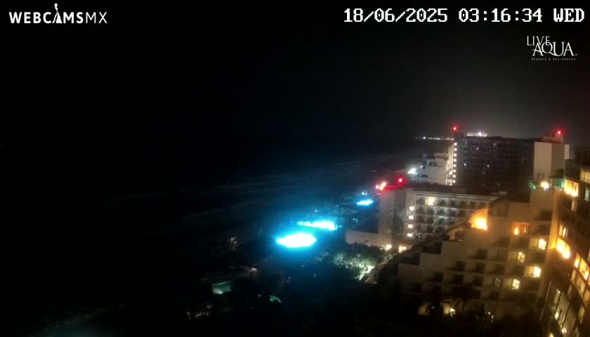 A nighttime view of a dark ocean, illuminated pools and brightly lit hotels along a coastline.
