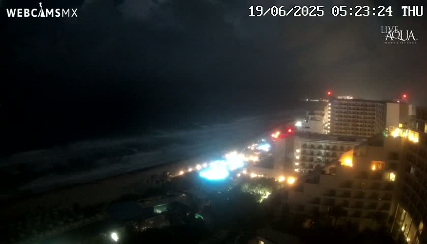 A nighttime view of a beach with dark, cloudy skies and illuminated hotels and streets along the shoreline.