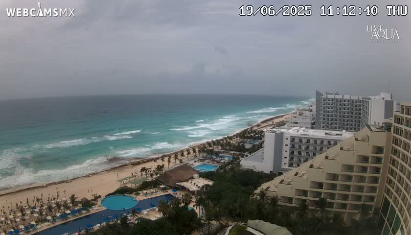 An ocean beach with turquoise water and waves under an overcast sky is viewed from above, showing resort hotels and swimming pools.