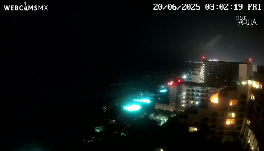 A nighttime view of illuminated hotel buildings along a dark ocean shore under a cloudy sky.