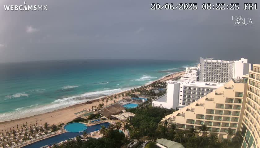An overcast sky overlooks a beach with turquoise water, waves breaking on the shore, and multiple resort hotels with swimming pools.