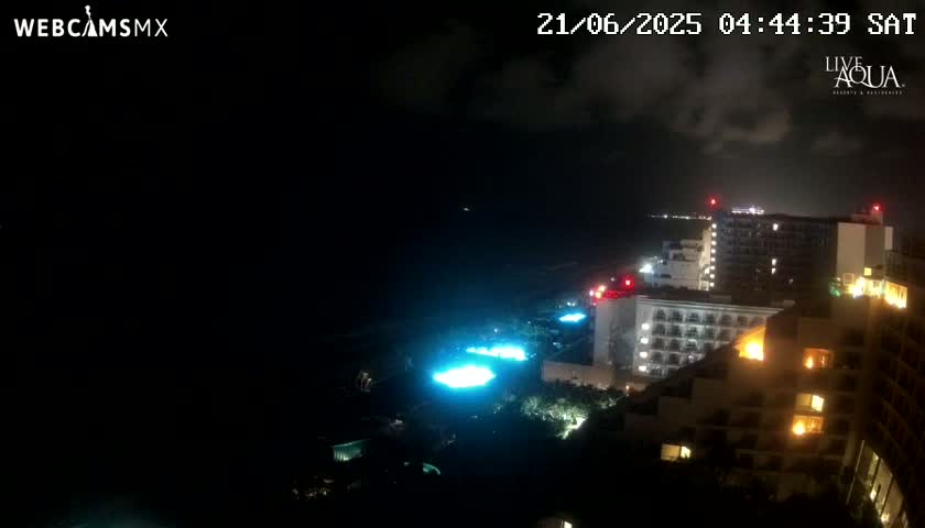A nighttime, coastal view of illuminated resort buildings, with a dark ocean and cloudy sky.