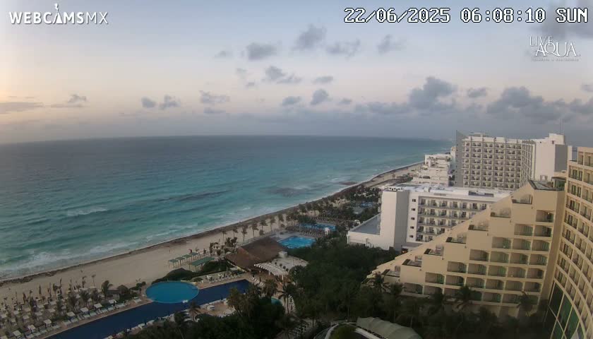 An aerial view shows a beach with turquoise water under a partly cloudy sky, with resort hotels and pools visible.
