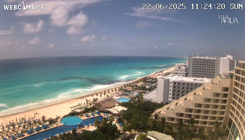 A sunny day shows a resort area with hotels, pools, and a long sandy beach bordering a turquoise ocean with small waves.
