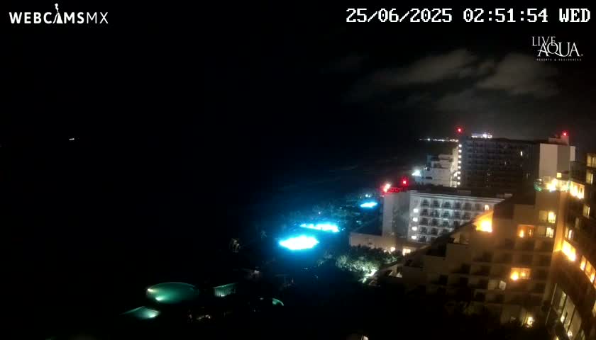 A nighttime view of illuminated hotels and pools near a dark ocean under a partly cloudy sky.