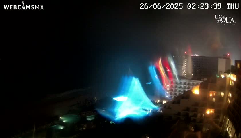 A nighttime view of a coastal city shows brightly lit buildings and a large, blue light display near the water under hazy conditions.