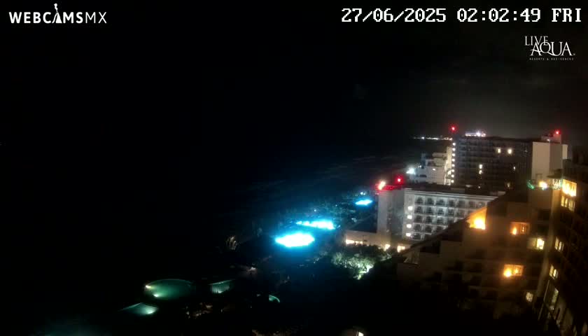 A nighttime view of illuminated resort buildings and pools near a dark ocean under a clear sky.