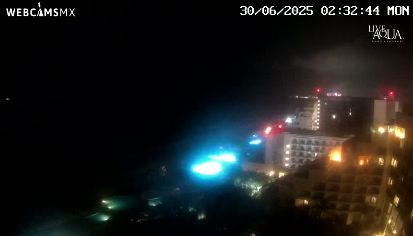 A nighttime, hazy view of illuminated buildings and pools near a dark body of water.