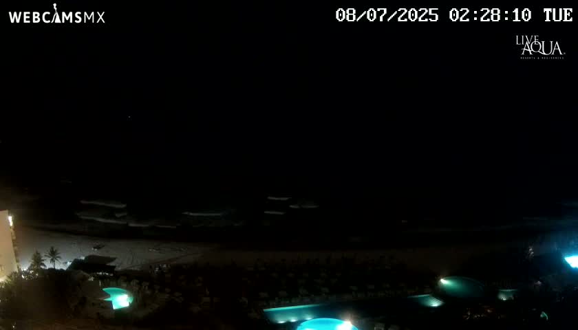 A nighttime view of a beach with calm waves and several illuminated pools at a resort.