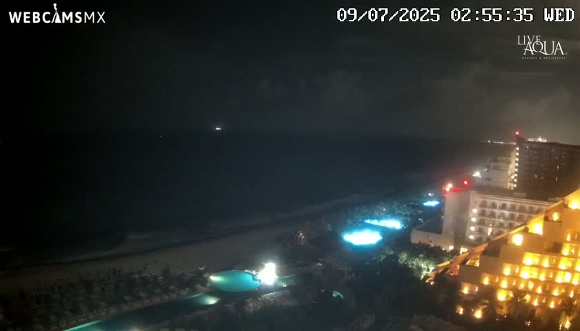 A nighttime view of a resort overlooking a dark ocean under cloudy skies shows illuminated pools and buildings.