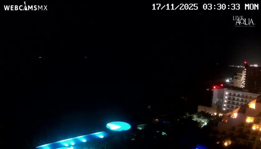 A high-angle view captures a brightly lit coastal resort with glowing blue pools and buildings next to a dark ocean under a clear night sky, with a distant coastline illuminated by lights.