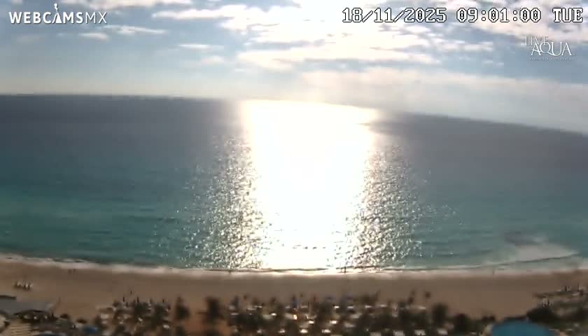A high-angle view captures a brightly lit coastal resort with glowing blue pools and buildings next to a dark ocean under a clear night sky, with a distant coastline illuminated by lights.