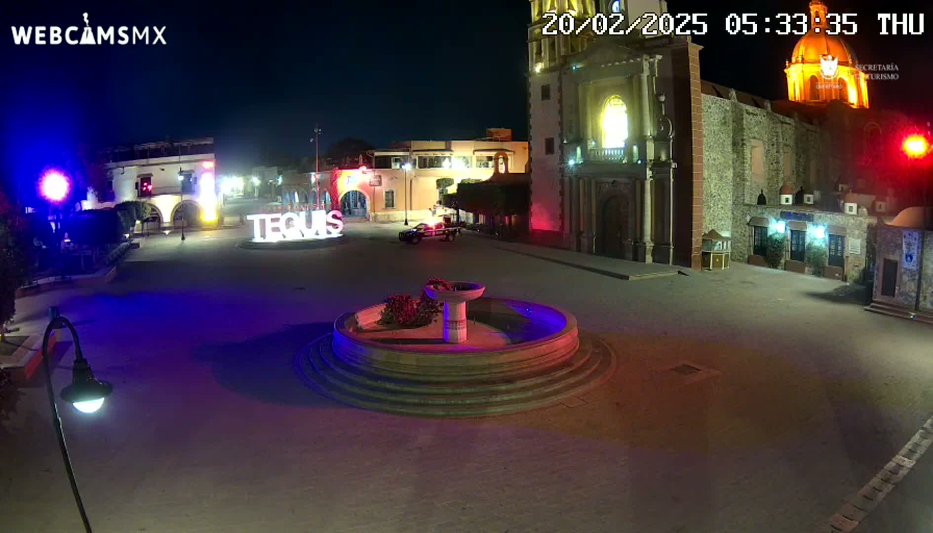 A nighttime view of a town square with a fountain in the center, illuminated buildings surrounding it, and a car parked nearby.