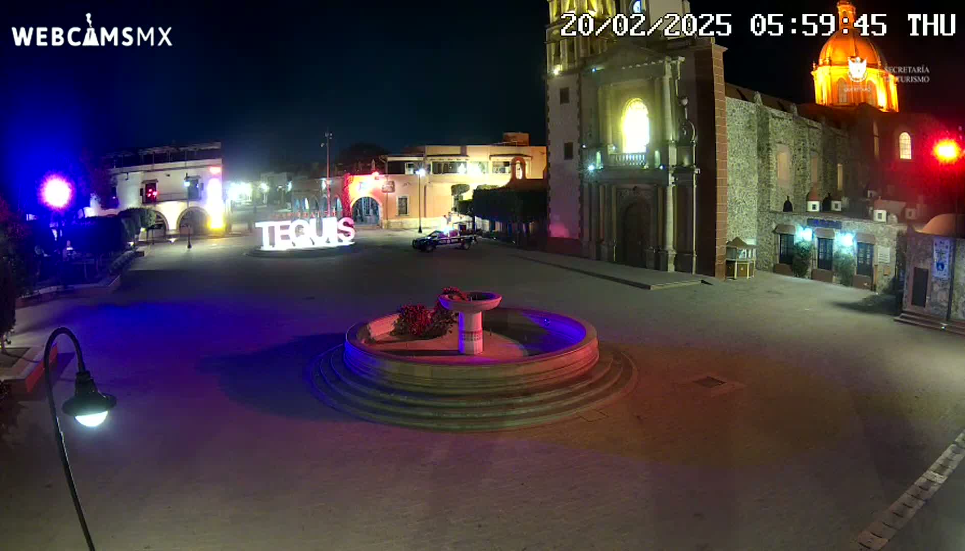 A town square at night, illuminated with colorful lights, features a circular fountain in the center, a church in the background, and a vehicle parked nearby.