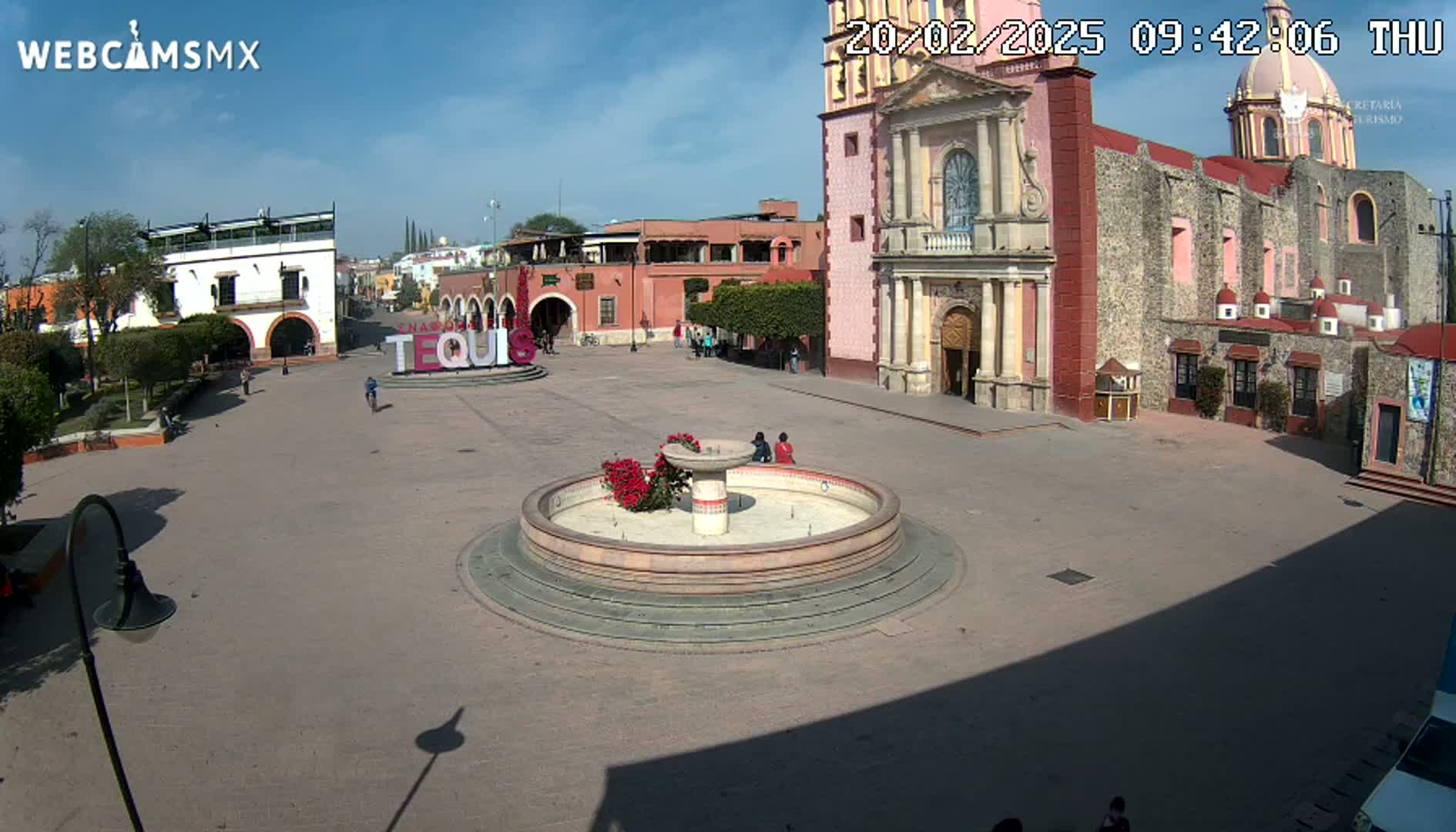 A town square with a fountain in the center, surrounded by colonial-style buildings under a sunny sky.