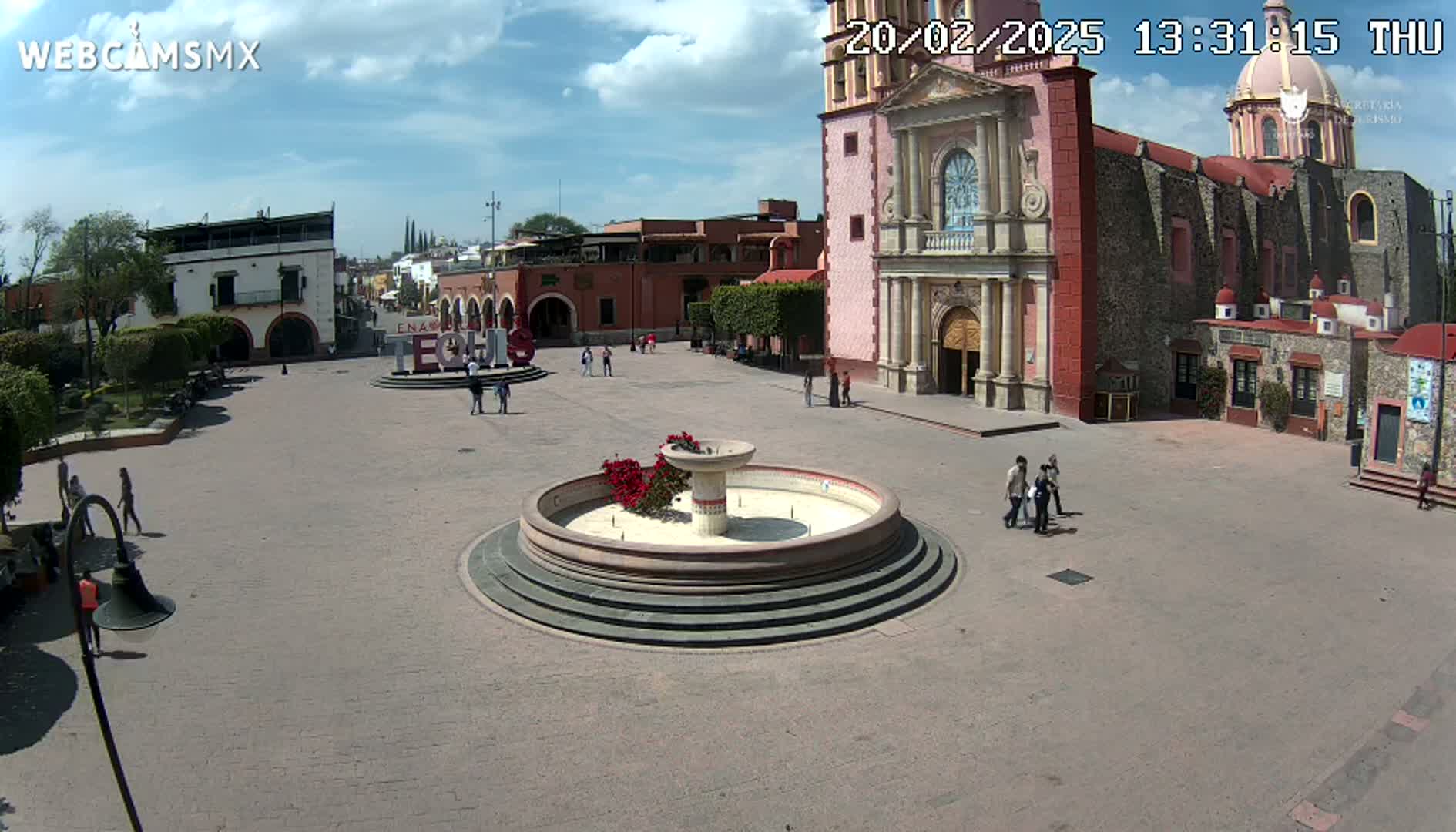 A town square with a fountain in the center, surrounded by colonial-style buildings, under a partly cloudy sky.