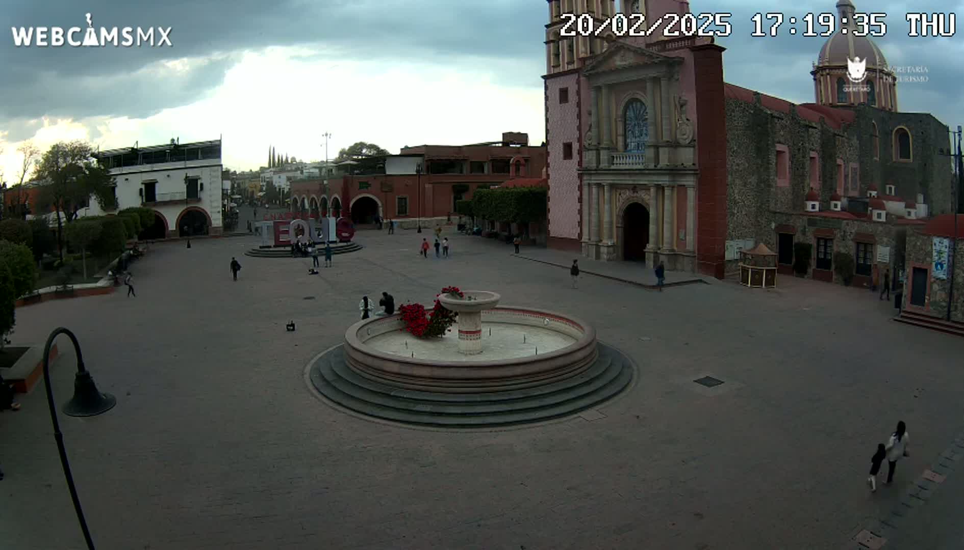 A town square with a fountain and a large church is mostly empty under a cloudy sky.