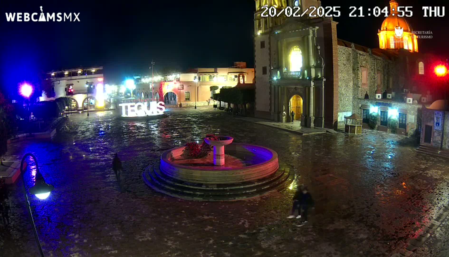 A wet town square at night, illuminated by lights, features a fountain and a large church in the background.