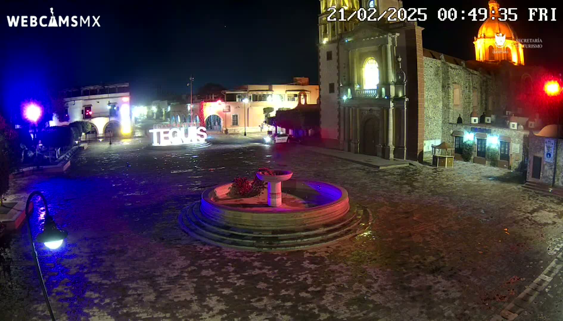 A nighttime view of a town square with a fountain, illuminated buildings, and wet pavement.
