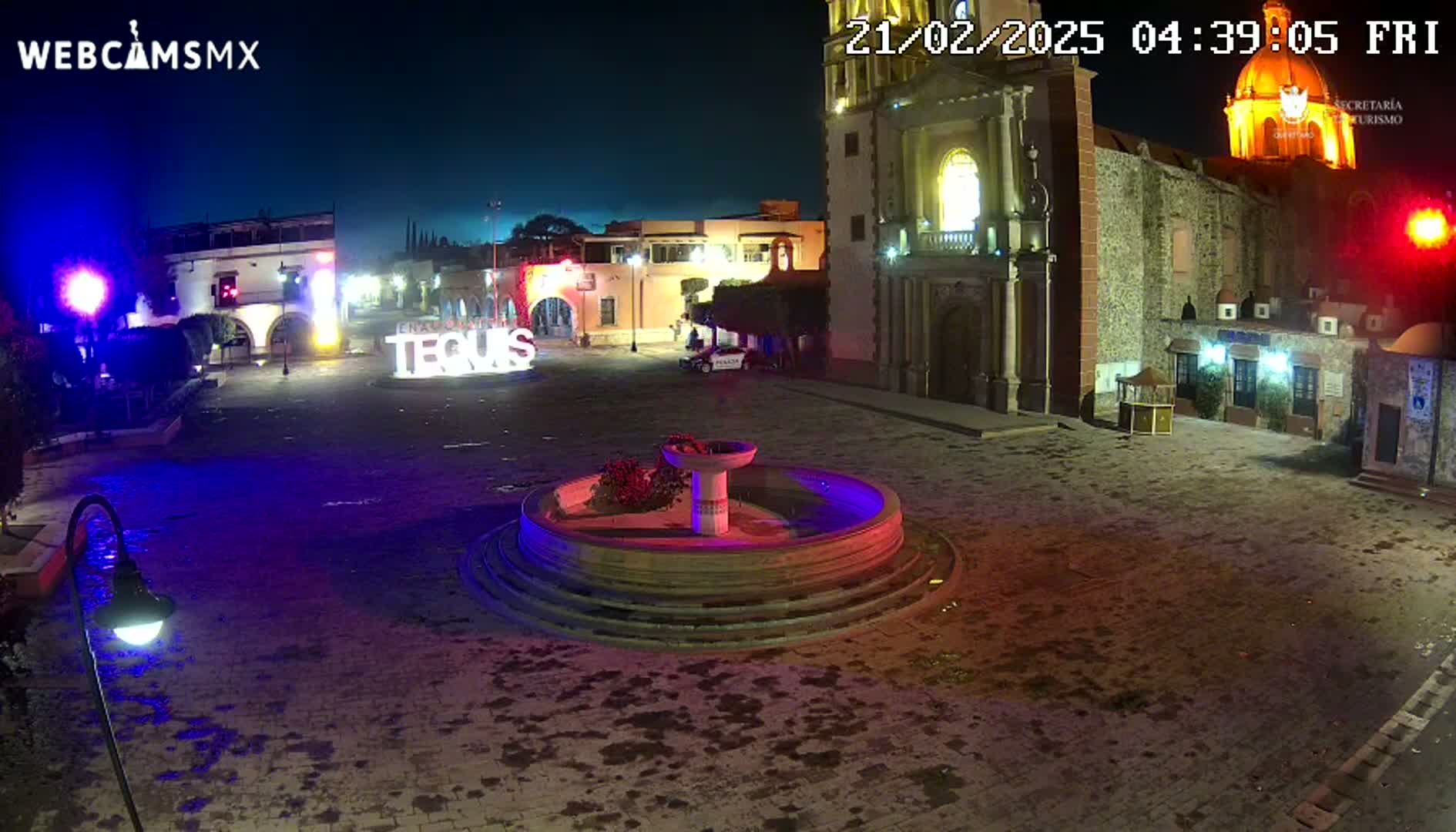 A nighttime view of a town square with a fountain, buildings, and decorative lights.