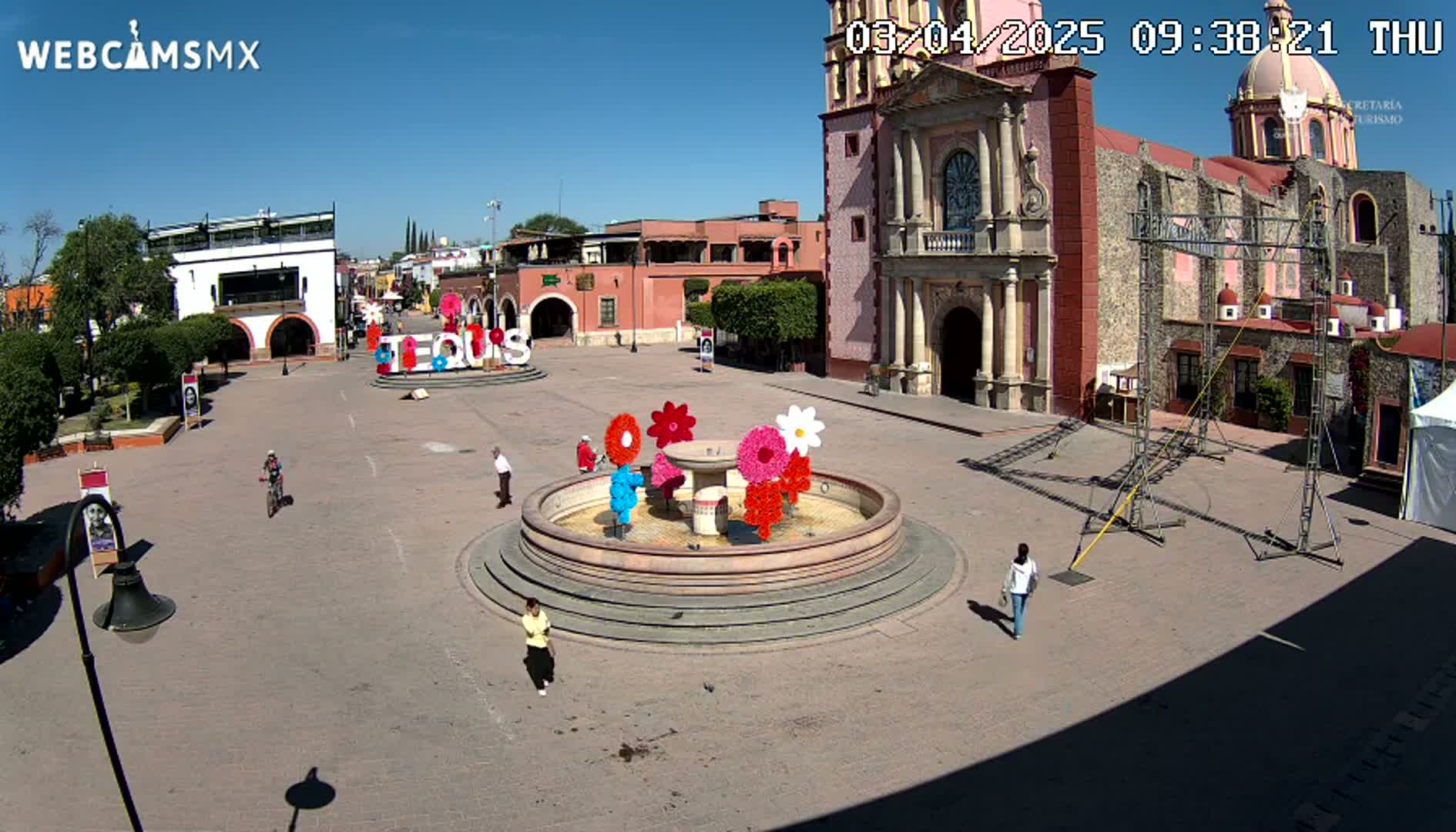 A town square with a fountain decorated with large colorful flowers, a church, and several people walking under a clear, sunny sky.