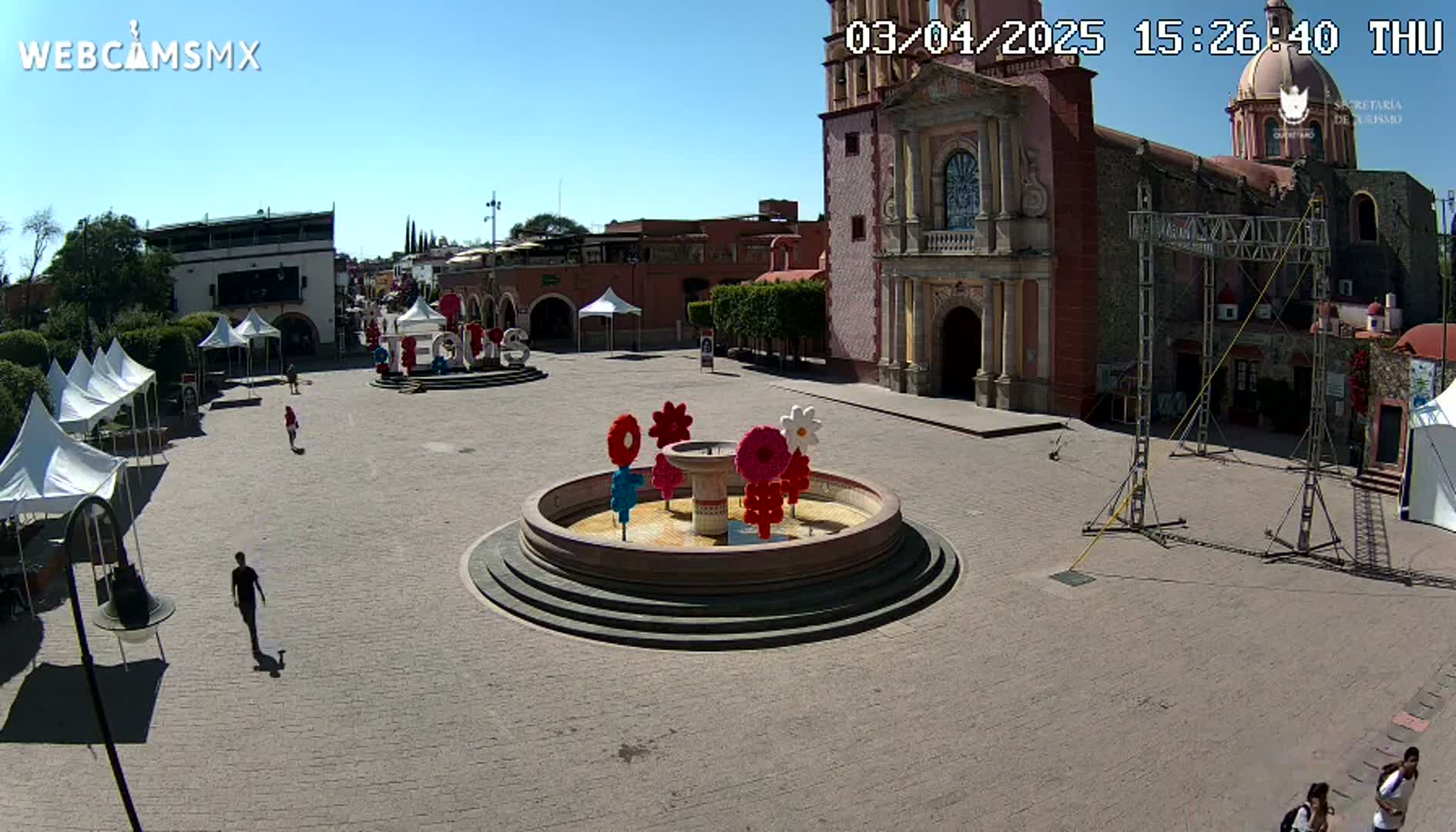 A town square with a fountain decorated with colorful flowers, several tents, and a large church under a clear, sunny sky.