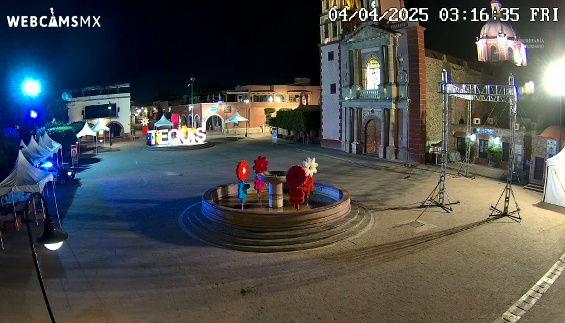 A nighttime view of a town square, featuring a fountain with colorful flower decorations, several tents, and a church in the background under dark skies.