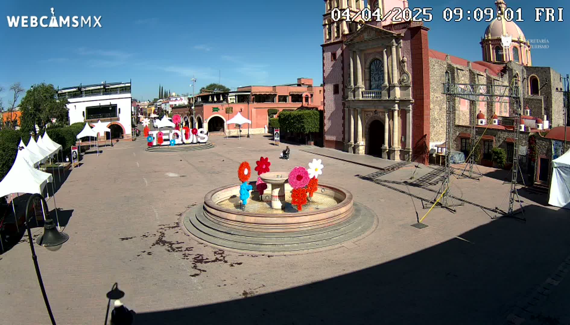 A town square on a sunny day features a fountain decorated with colorful flowers, several white tents, and a large church in the background.