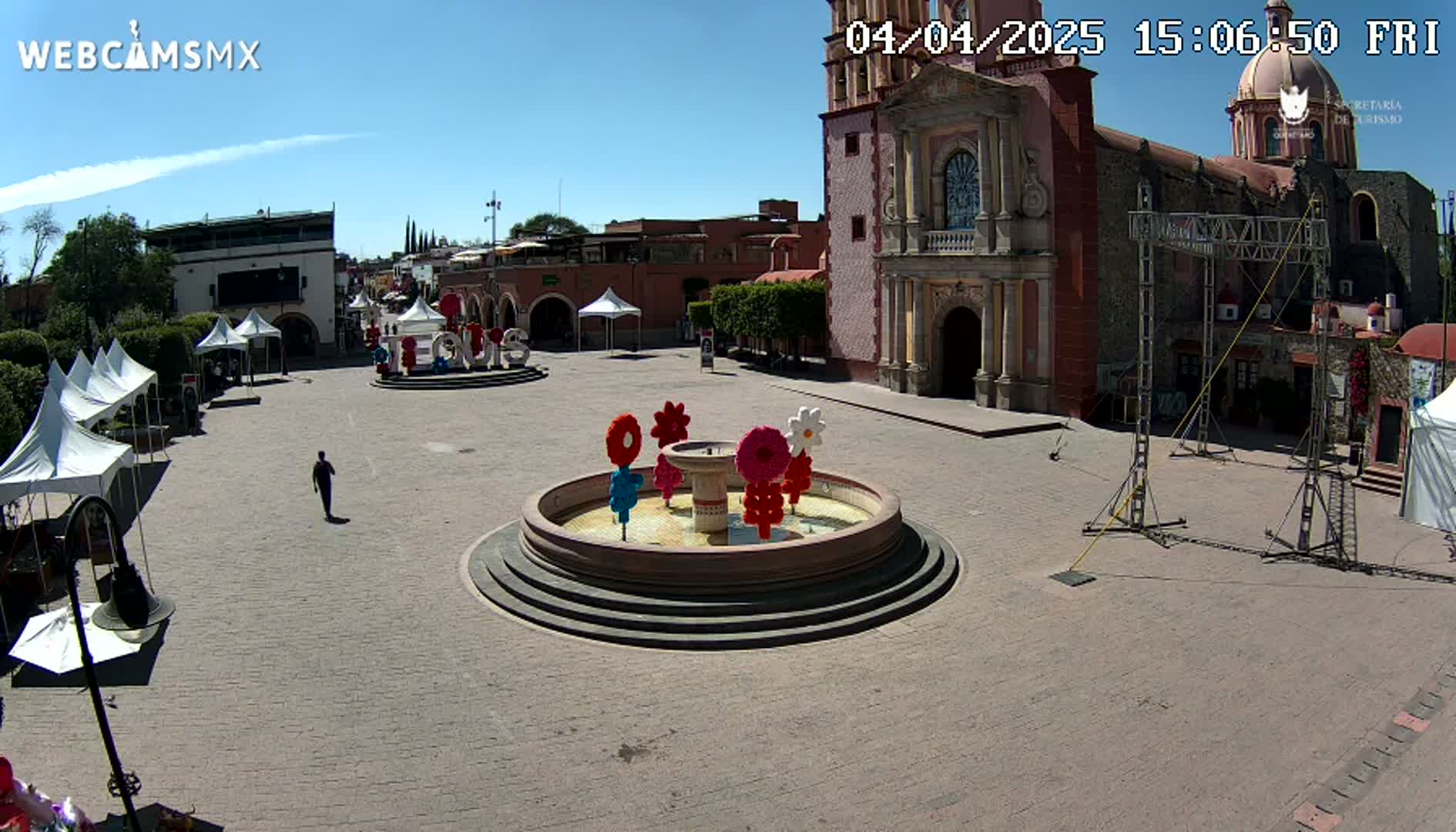 A town square under a clear blue sky features a central fountain decorated with colorful flowers, surrounded by buildings, including a church, and several white tents.
