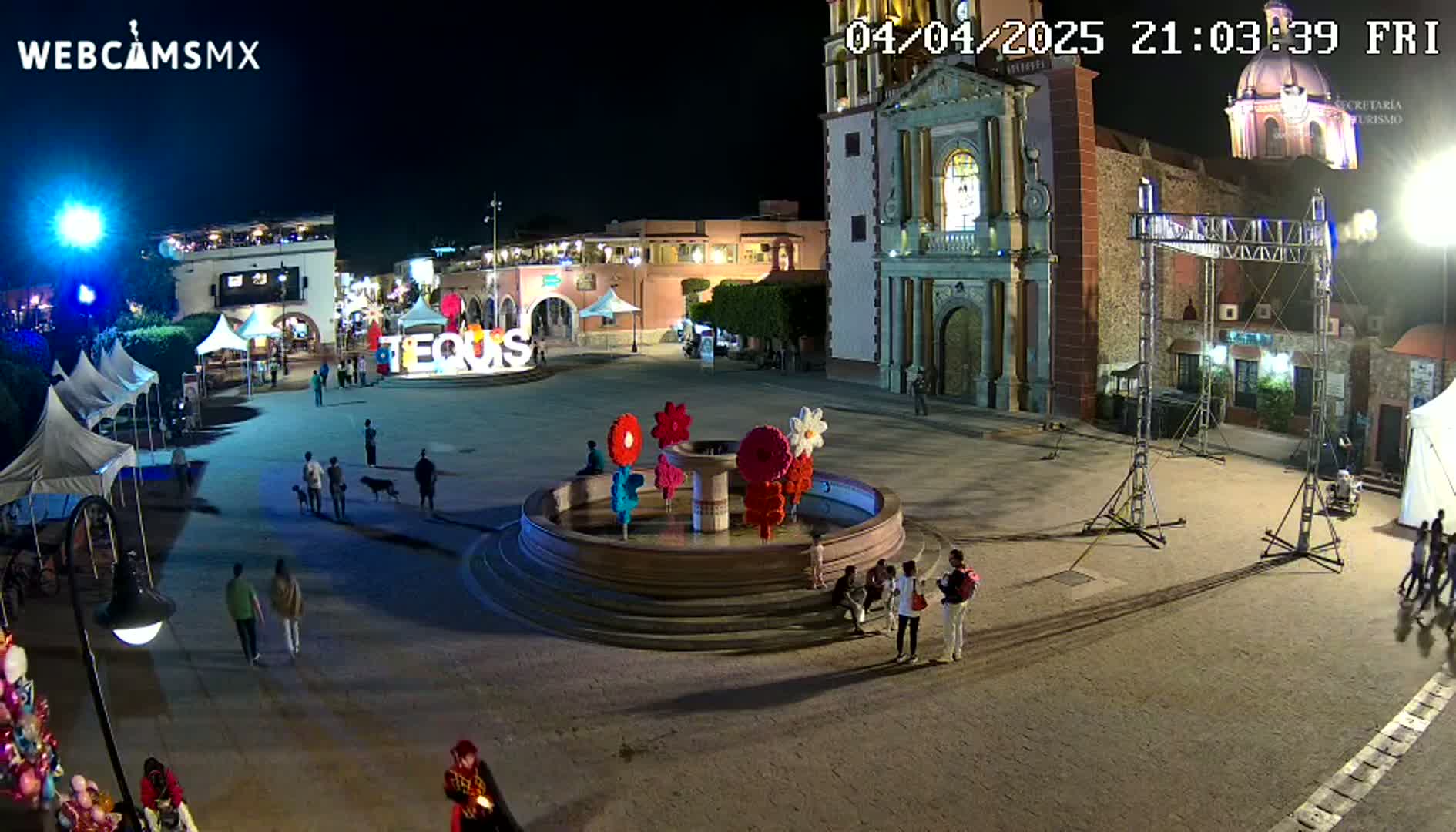 A town square at night, illuminated, features a fountain decorated with large colorful flowers, surrounded by people, buildings, and temporary event structures.