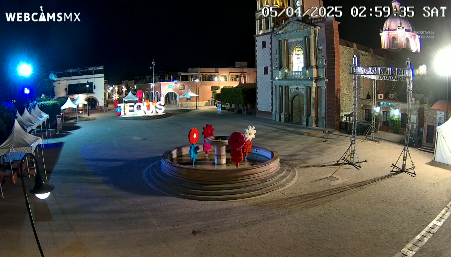 A town square at night, illuminated, features a circular fountain decorated with colorful flowers, several tents, a church, and stage equipment.