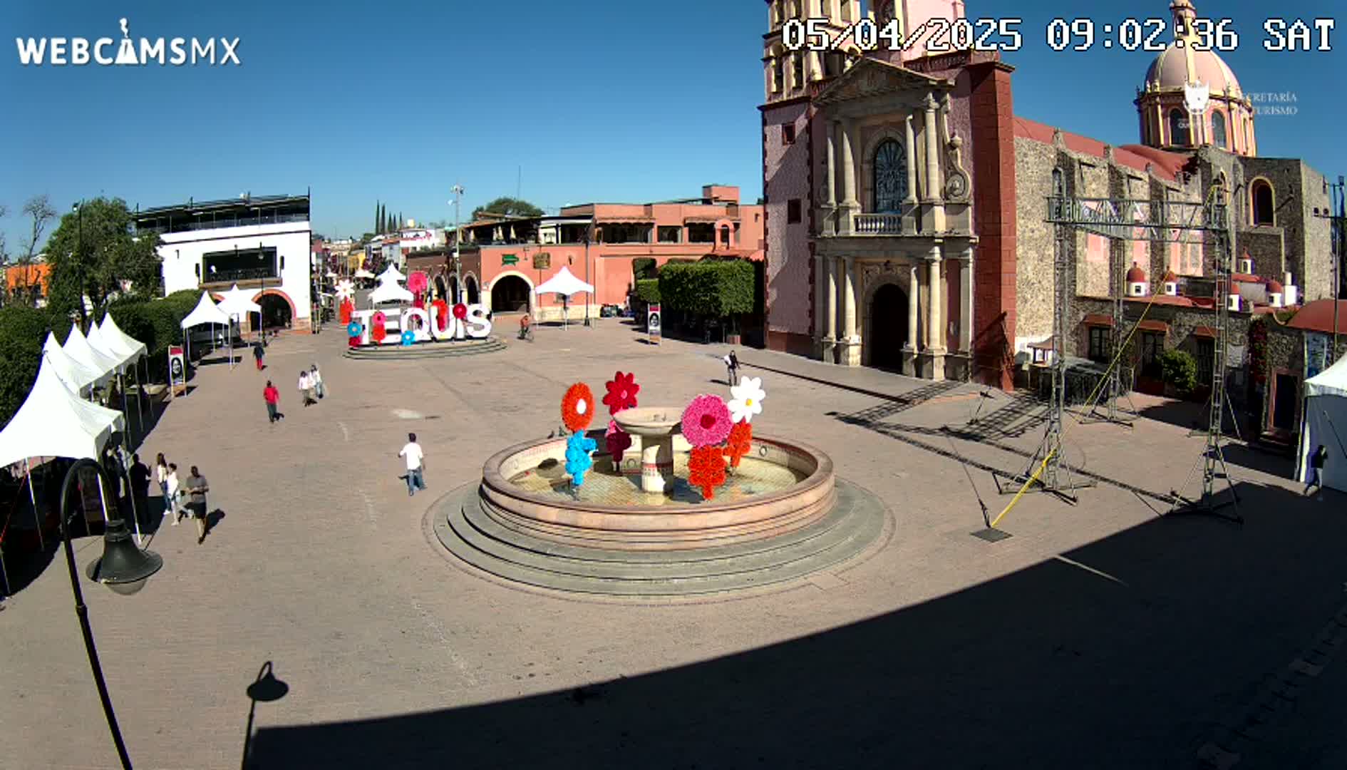 A town square on a sunny day features a fountain decorated with large artificial flowers, several white tents, a church, and a few pedestrians.