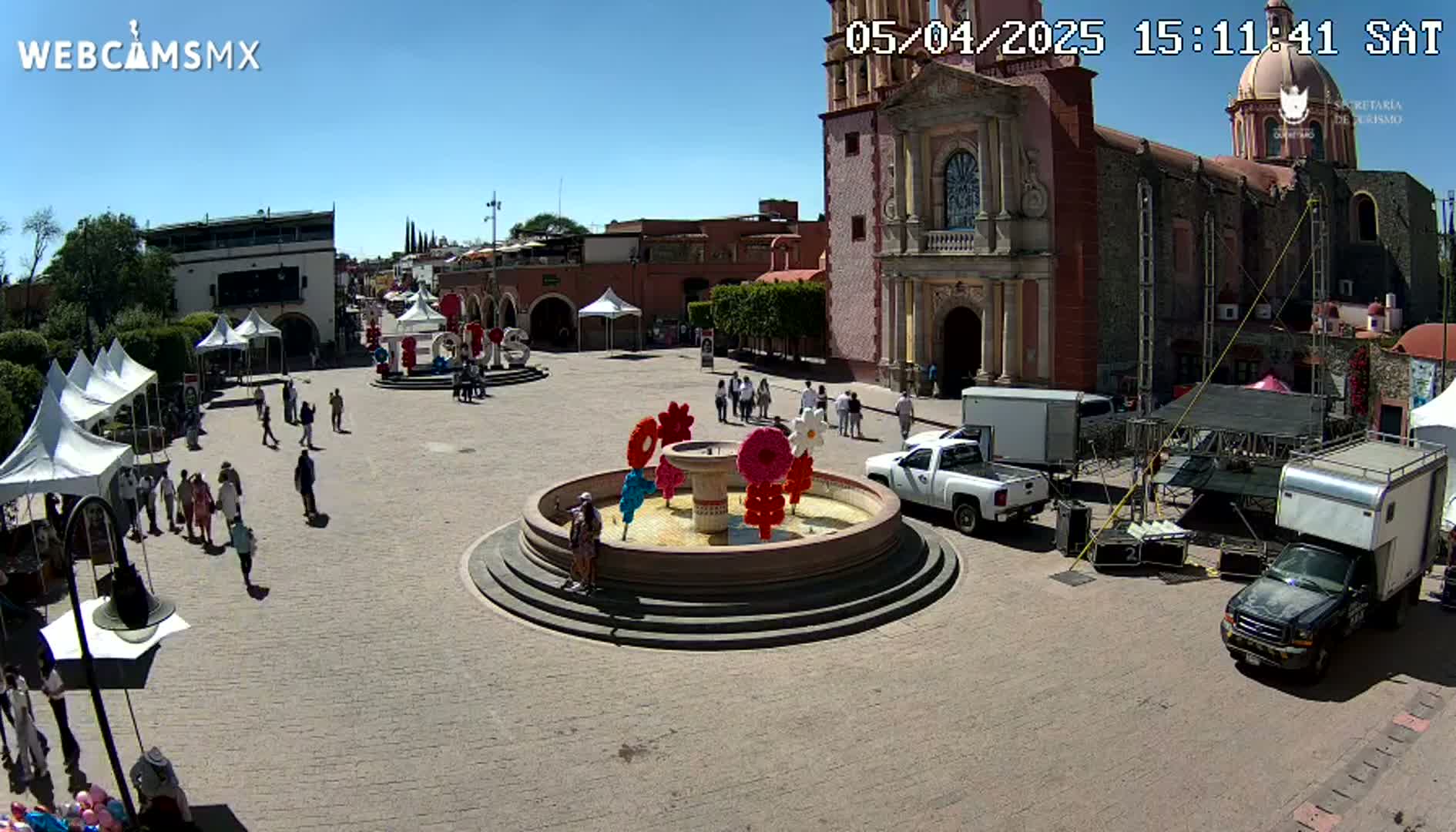 A town square with a fountain decorated with colorful flowers, several tents, people walking around, and a large church in the background on a sunny day.
