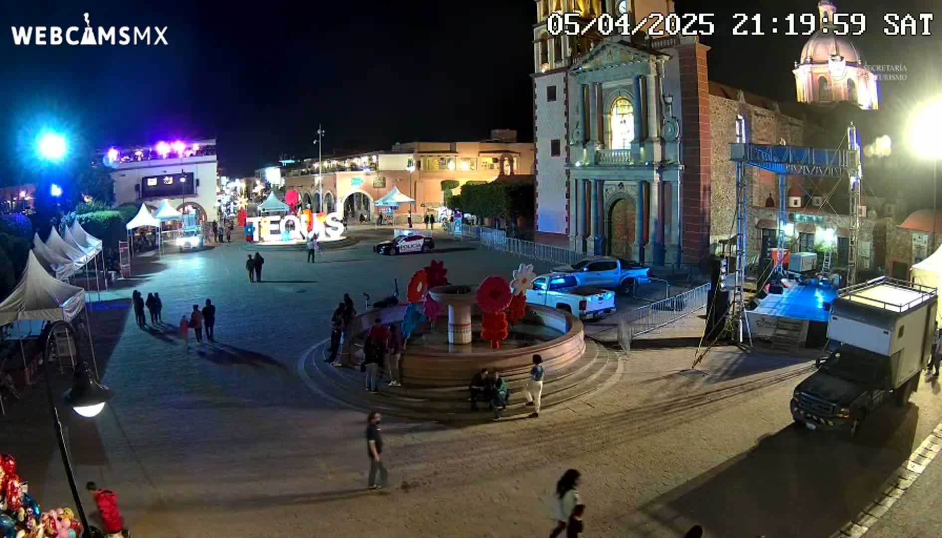 A nighttime town square with a fountain, several people, tents, vehicles, and a church in the background.