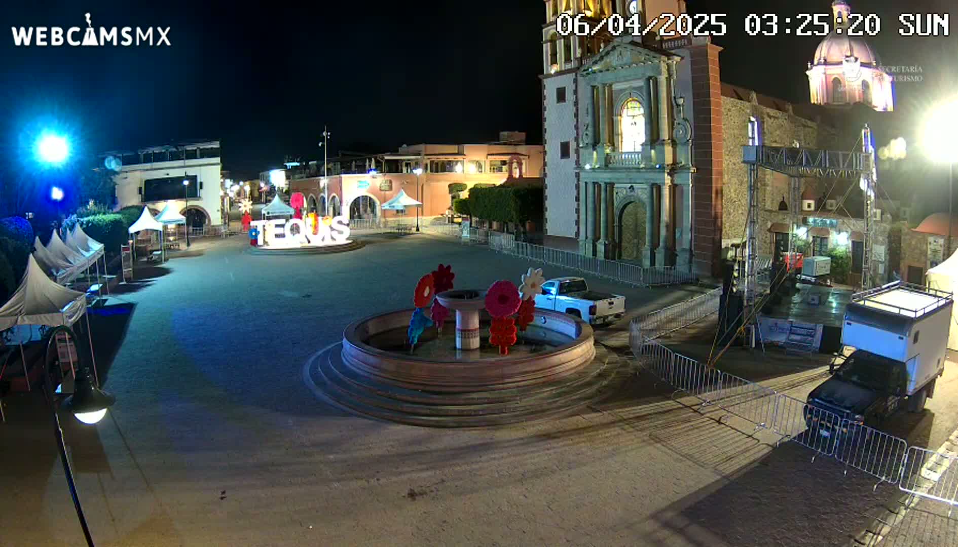 A town square at night, illuminated by artificial light, features a fountain with colorful floral decorations, several tents, a church, and vehicles parked nearby.