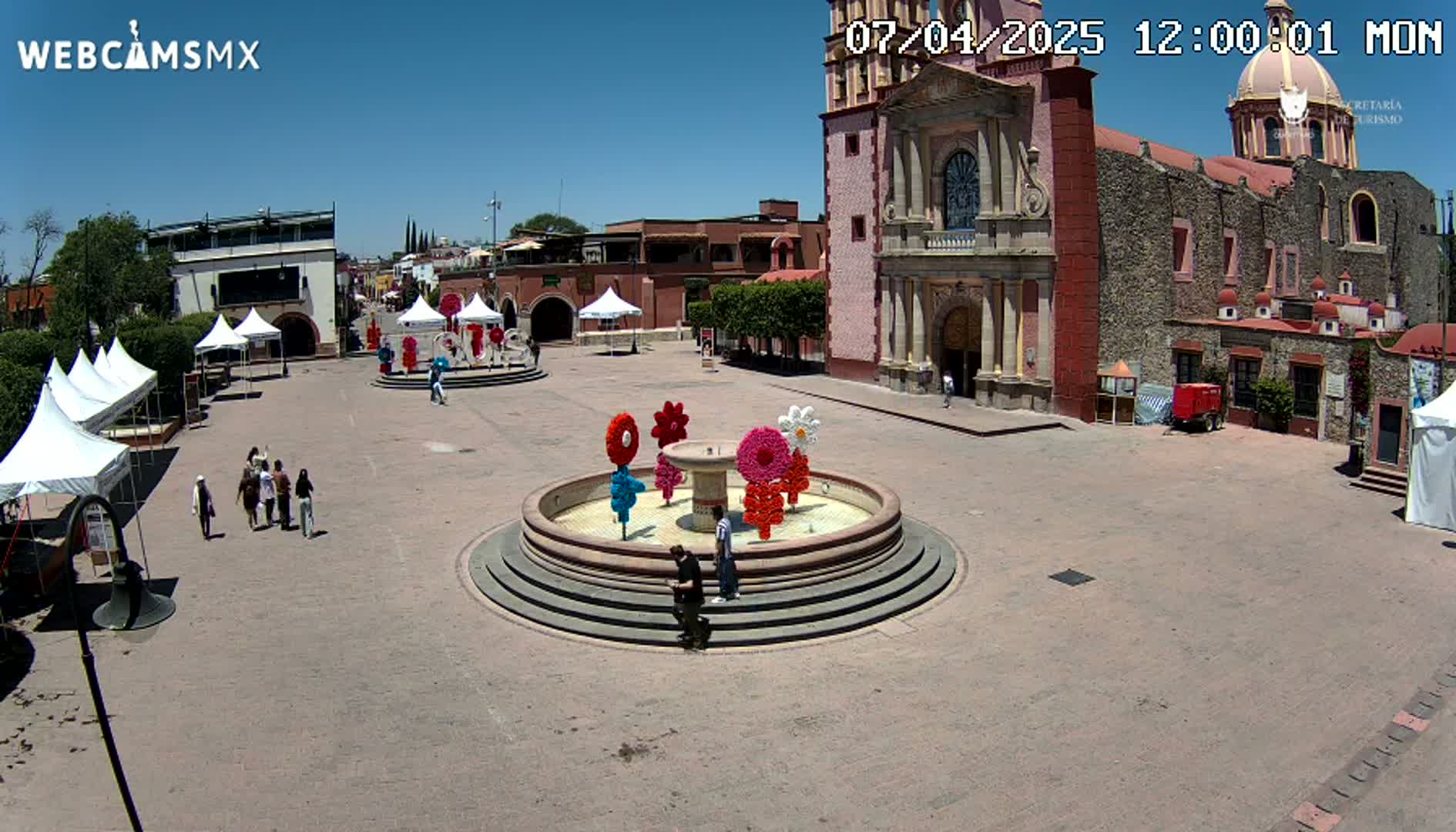 A town square on a sunny day features a fountain decorated with colorful flower designs, several white tents, a church, and a few people walking around.