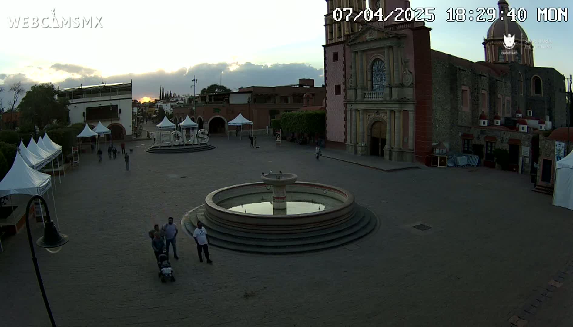 A town square at sunset features a central fountain, several people, market tents, and a large church in the background.