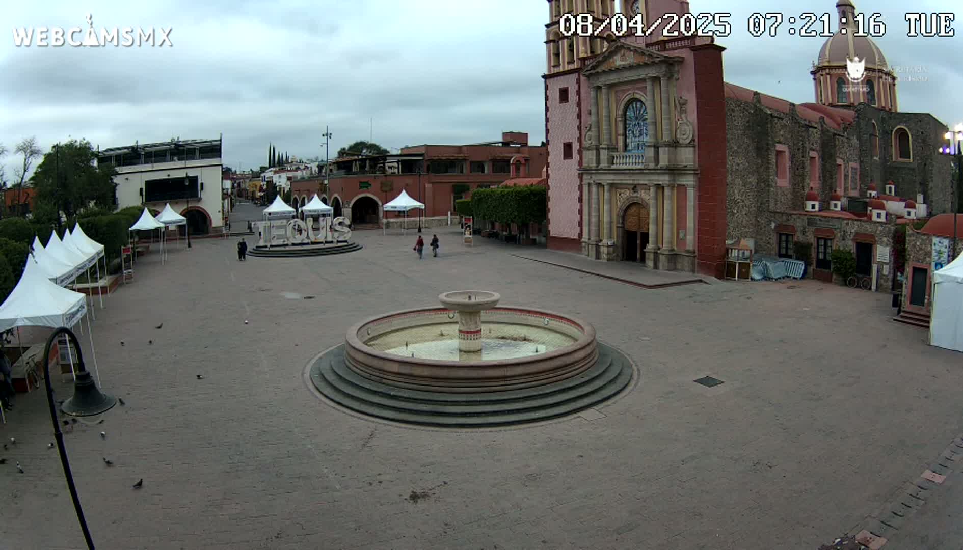 A town square with a fountain, several white tents, and a large church in the background under an overcast sky.