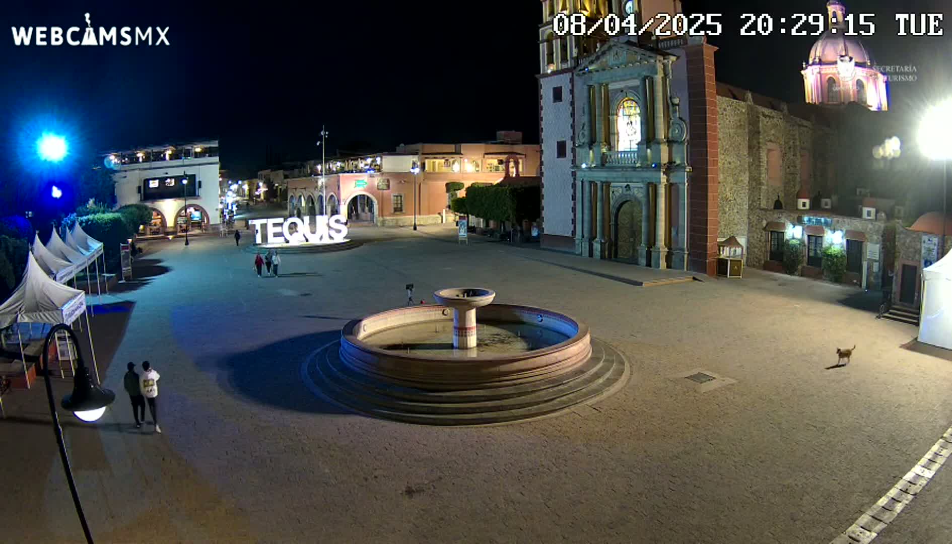 A nighttime view of a town square with a fountain, several buildings, and a few people, under clear skies.