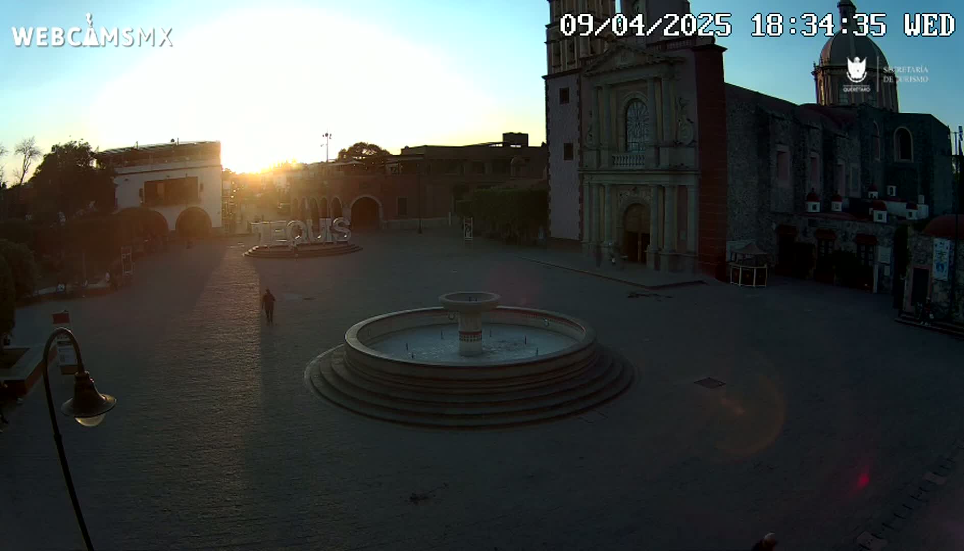 A town square at sunset, featuring a fountain and a large church in the background, under clear skies.