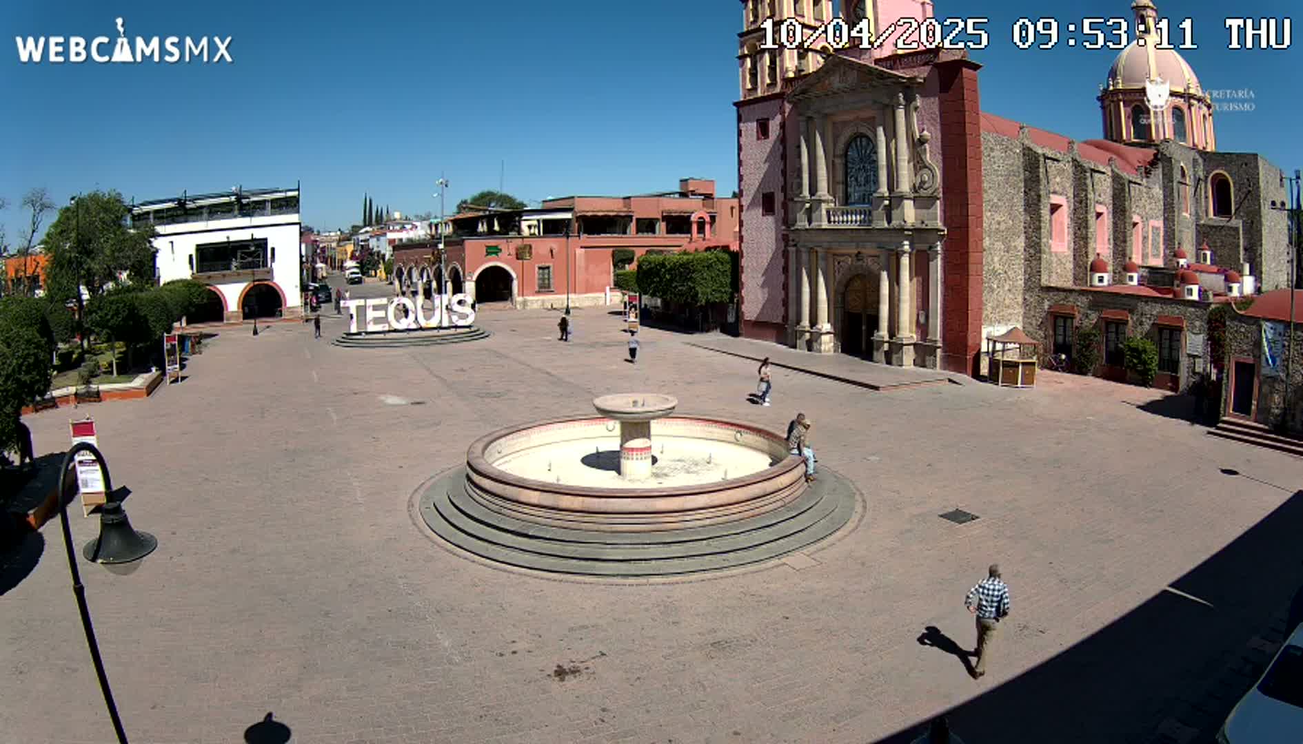 A town square with a fountain, several buildings including a large church, and a few people are visible under sunny skies.