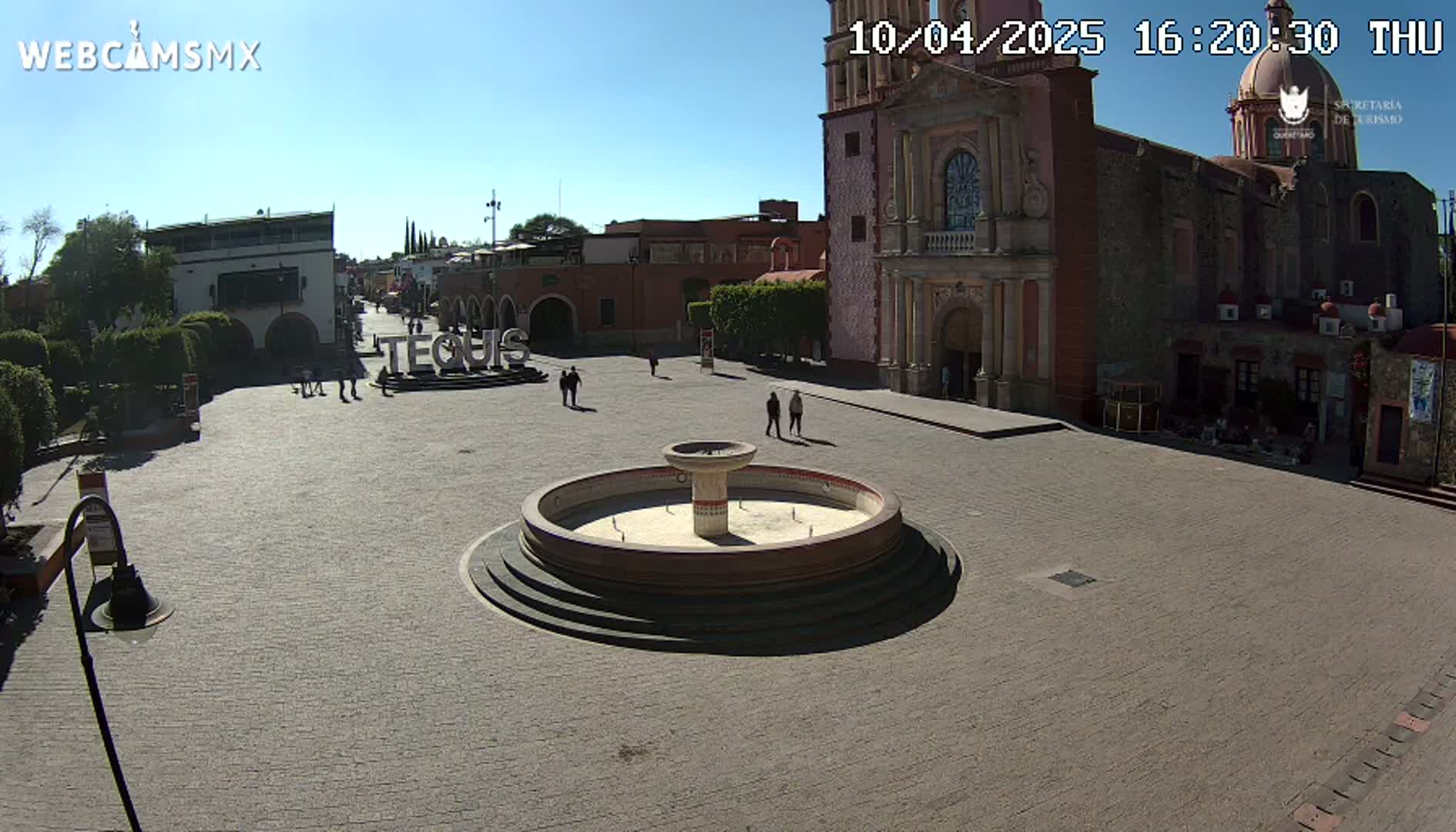 A town square with a central fountain is shown under sunny skies, adjacent to a large church and other buildings.