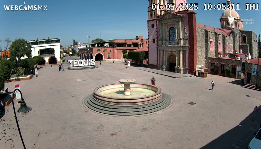 A town square with a fountain in the center, surrounded by buildings including a large church, under a clear blue sky.