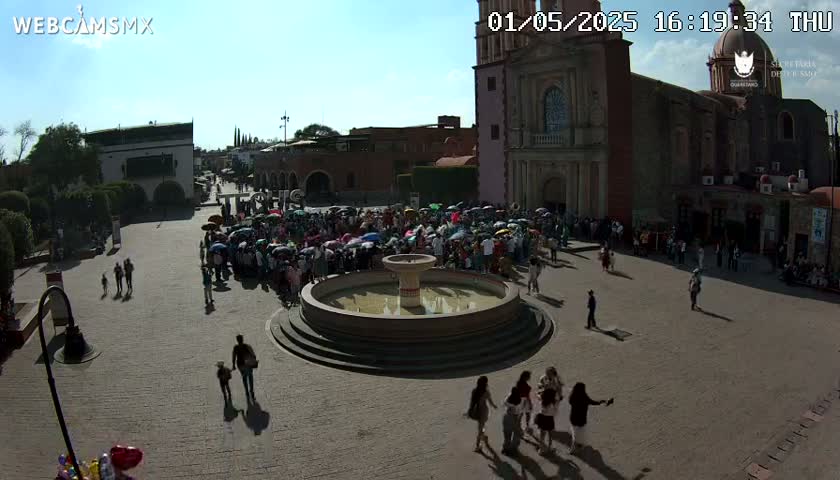 A large crowd of people gathers around a fountain in a town square on a sunny day, in front of a large church.