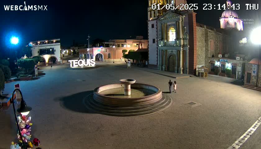 A nighttime view of a town square with a fountain, buildings, and a couple standing near it.